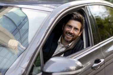 Pahalı bir arabanın direksiyonunda oturan ve camdan dışarı bakıp kameraya gülümseyen yakışıklı ve başarılı bir iş adamının yakın plan portresi..