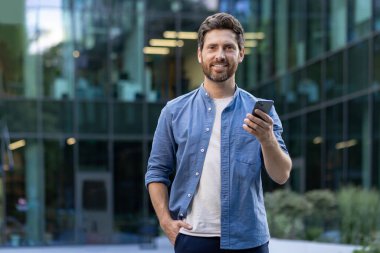Sıradan giyinmiş, sakallı, telefon kullanan, güneşli bir günde cam binanın yanında duran, kendine güveni tam bir iş adamı..