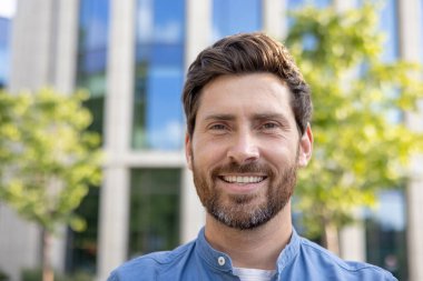 Sakallı neşeli iş adamı dışarıda eğleniyor. Arka planda ofis binaları olan günlük kıyafetli bir adam..