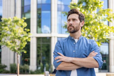 Sıradan tişörtlü, kollarını kavuşturmuş, bir ofis binasının dışında düşünceli görünen olgun sakallı bir adam..