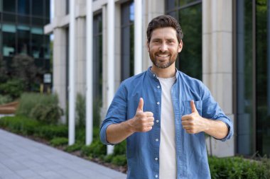 Günlük kıyafetlerle gülümseyen sakallı iş adamı, bir şirket binasının önünde durup başarıyı işaret ediyor..