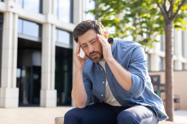 Sıkıntılı, olgun iş adamı baş ağrısı yüzünden tapınakları ovalıyor, ofis binasında oturuyor, iş stresi ve tükenmişlik hissi içinde..