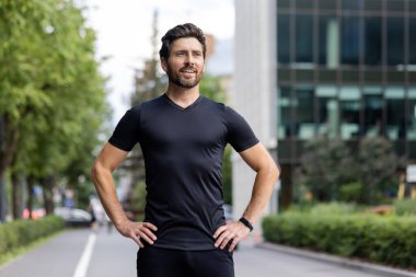 Gülümseyen ve kendine güveni tam bir sporcunun portresi bir şehir caddesinde duruyor, ellerini yan yana tutuyor ve yan tarafa bakıyor..