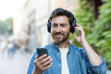 Sakallı neşeli, olgun bir adam kulaklıkla müzik dinlerken cep telefonu kullanıyor, şehir ortamında yürüyor..