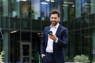 Sakallı, olgun bir işadamı modern bir ofis binasının önünde akıllı telefonunu kullanırken gülümsüyor..