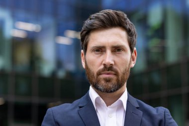 Seçkin, olgun, sakallı, takım elbiseli bir iş adamının portresi. Bir ofis binasının önünde duruyor..