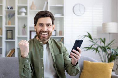 Sakallı, mutlu, olgun bir adam bir telefon tutarken heyecanlı bir şekilde yumruğunu sıkar, evdeki kanepede oturur ve başarısını gösterir..