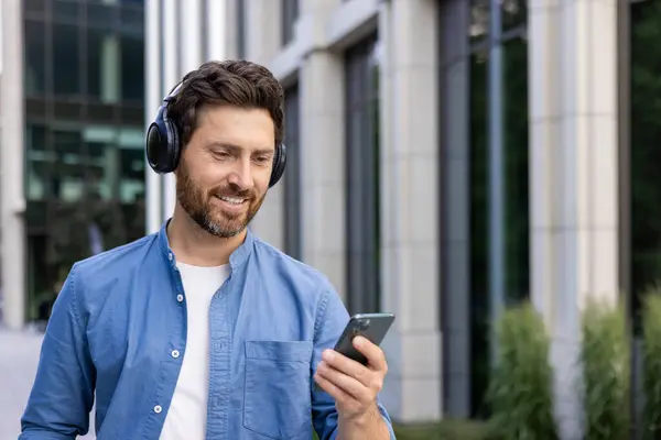 Close-up photo of a young smiling man standing on a city street wearing headphones and using a mobile phone. Listens to music, watches videos, talks on a video call.