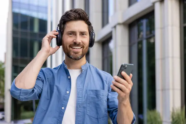 Mature businessman in casual attire enjoys music on headphones while using a smartphone outside an office building.
