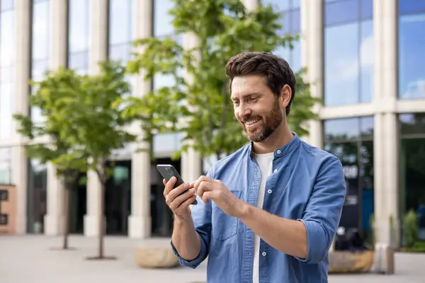 Casual mature businessman with a beard happily uses a phone outside modern office buildings, signifying work flexibility.