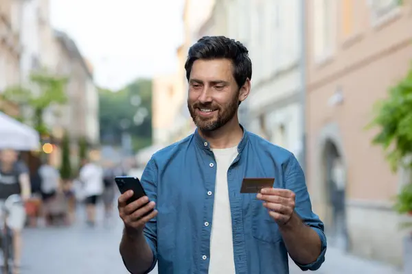 Smiling bearded man in casual attire using a smartphone and holding a credit card on a bustling city street in the evening.