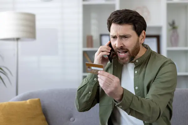 Anxious mature man with beard on phone call holding credit card, sitting on sofa in a cozy living room apartment.
