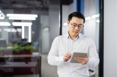 Elinde tablet bilgisayarı olan ciddi düşünen bir adam, ofisin içinde Asyalı bir adam, tişört okuyan ve internetten veri analiz eden bir iş adamı..