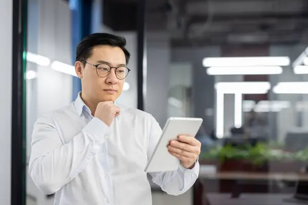 Serious thinking man with tablet computer in hands, asian man at workplace inside office, businessman in shirt reading and analyzing online data using app.