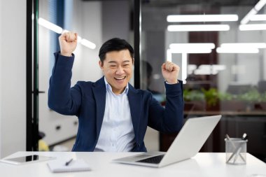 İş yerinde dans eden neşeli bir adam, iş yerinde takım elbiseli başarılı Asyalı bir işadamı ofis içinde dizüstü bilgisayarla oturmuş başarılı başarıları kutluyor, mutlu bir gülümseme..