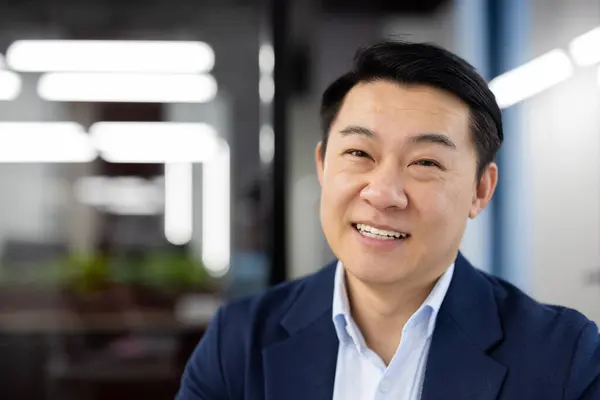 Close-up portrait of Asian man inside office, businessman successful smiling looking at camera, boss in business suit at workplace, successful investor financier, banker.