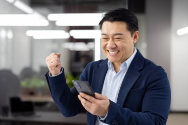 Ofis içinde telefonu olan Asyalı bir adam başarılı başarıları kutlayan bir işyeri, iş adamı internetten iyi haberler aldı, olgun başarılı bir patron gülümsüyor. .