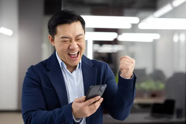 Asian man with phone in hands inside office an workplace celebrating successful achievement results, businessman received notification of good news online, mature successful boss smiling reading .