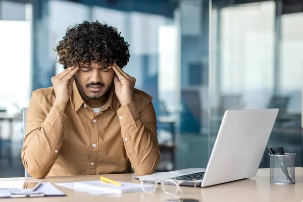 Tired and sick young Indian man sitting in the office at the desk, and grimacing, suffering from a severe headache and pressure, massaging his head with his hands.