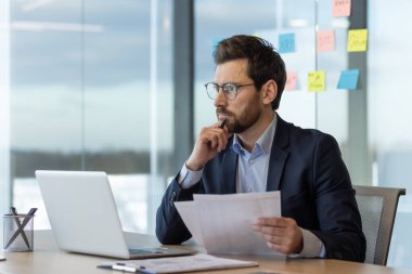 Sakallı, olgun bir işadamı, kağıtları dikkatle inceleyip, etrafı teknoloji ve yapışkan notlarla çevrili..