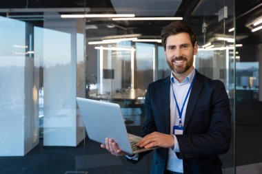 Çağdaş ofis ortamında elinde dizüstü bilgisayarla gülümseyen olgun iş adamı liderlik ve profesyonelliği yansıtıyor..