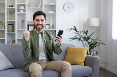 Sakallı neşeli, olgun bir adam kazanan bir jest yapıyor. İyi aydınlatılmış bir oturma odasındaki kanepede rahatça otururken telefonu tutuyor..