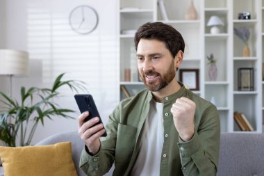 Sakallı, olgun bir adam akıllı telefonunu koltukta kullanırken neşe gösteriyor. Başarı ve mutluluk modern oturma odasında resmedildi..