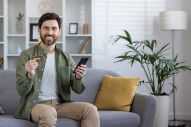 Sakallı, mutlu ve olgun bir adam. Elinde bir kredi kartı ve akıllı telefon var. Parlak oturma odasından online alışverişler için hazır..