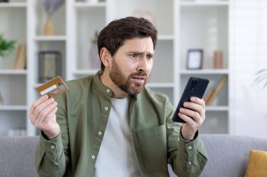 Sakallı, olgun bir adam elinde kredi kartı ve akıllı telefon varken endişeli görünüyor. Mali sorunlarla karşı karşıya olabilir..