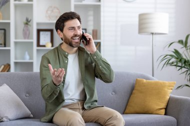 Oturma odasında rahat bir kanepede oturup, mutluluk ve rahatlığı ifade ederken, sakallı, gülümseyen yetişkin bir adam telefonda konuşuyor..