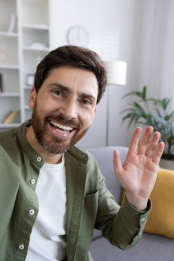 Sıradan gömlekli neşeli sakallı bir adam kameraya el sallıyor. Evdeki rahat bir kanepede otururken arkadaşça bir hava yayıyor..