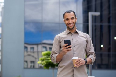 Bir kişi elinde bir akıllı telefon ve bir kahve fincanı tutuyor, arka planda ağaçlar ve binalarla dışarıda duruyor.