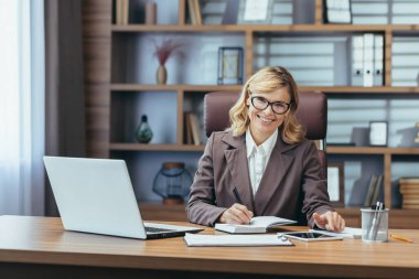 Modern bir ofisteki neşeli, olgun bir iş kadını masasında dizüstü bilgisayar ve evrak işleriyle meşgul bir şekilde çalışıyor..