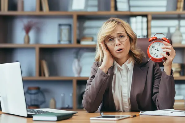 An overwhelmed senior businesswoman holds a red alarm clock, expressing urgency and stress at her office desk surrounded by work.