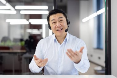 Bir ofis ortamında etkin bir şekilde konuşan, kulaklık takmış neşeli bir Asyalı işadamı..