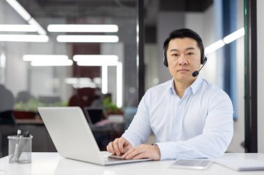 Güncel ofis ortamında kulaklığı olan, masa başında dizüstü bilgisayar kullanan aklı başında Asyalı bir adam..