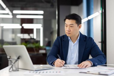 Profesyonel Asyalı işadamı iyi aydınlatılmış modern bir ofiste dizüstü bilgisayarla belgelere bakıyor..