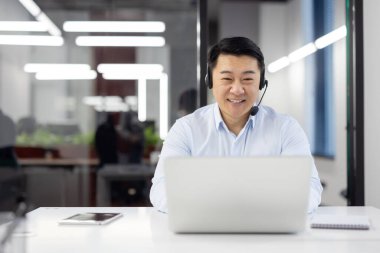 Ofisin içinde kulaklıklı bir adam, gömlek giyen Asyalı bir müşteri hizmetleri çalışanı, video görüşmesi için laptop kullanan bir iş adamı ve müşteri danışmanlığı, telefon satışları.,