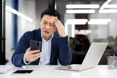İş yerinde üzgün Asyalı bir adam, iş elbisesi içinde hoşnutsuz bir çalışan, elinde telefon, iş adamı akıllı telefondan kötü haberlerle internetten bilgilendirme mesajı aldı, patron şaşırdı..