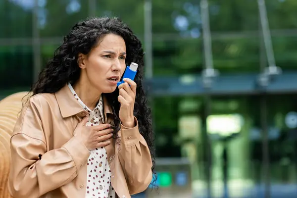 Concerned young Hispanic businesswoman using an asthma inhaler during a break, with an office building in the background.