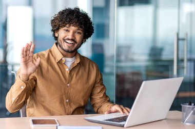 Ofiste çalışan mutlu bir Hintli gencin portresi. Kameraya bakar, selamlar ve dalgalar.