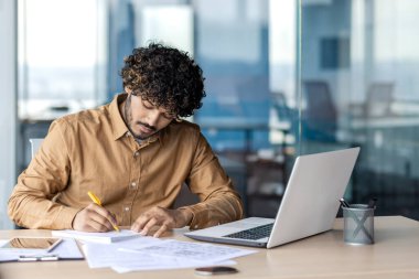 Genç Hintli erkek işadamı ofiste çalışıyor, dizüstü bilgisayarla masada oturuyor ve belgeler ve faturalar yazıyor..