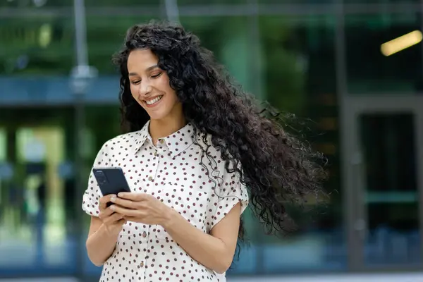 Young beautiful Latin American woman walking in the city with phone in hands, business woman after working day, smiling with satisfaction, student using application on smartphone.