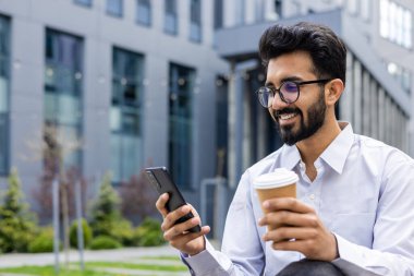 Elinde telefon ve bir bardak sıcak içecekle genç bir adam, iş adamı mutlu bir şekilde sosyal ağlara bakıyor, ofis binasının dışında dinleniyor, akıllı telefon kullanıyor..