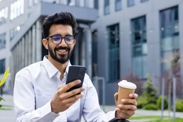 Young man with phone in hands and a cup of hot drink close up, businessman smiling contentedly browsing social networks, worker resting outside office building, using smartphone.