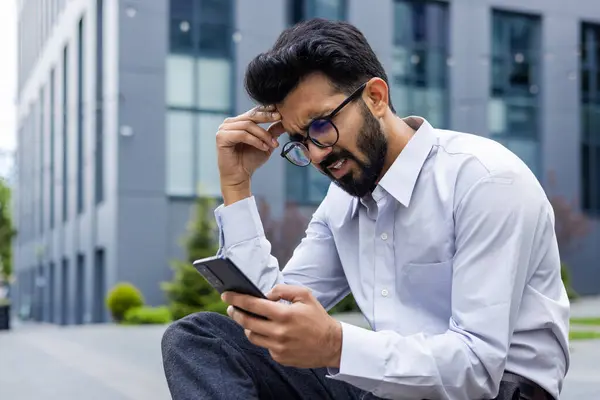 Young man depressed, sad dissatisfied and unhappy outside office building, holding phone, reading bad news from smartphone, businessman in shirt after work.