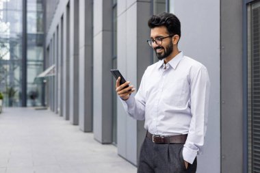 Çağdaş ofis mimarisinin dışında akıllı telefon kullanarak iş kıyafetleri içinde gülümseyen genç adam güven ve bağlantı gösteriyor..
