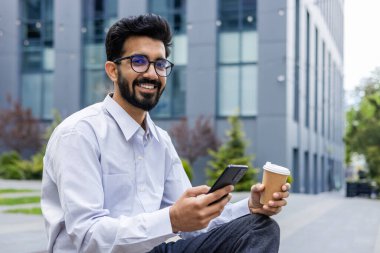Modern ofis binalarının önünde cep telefonu ve kahveyle gülümseyen beyaz gömlekli profesyonel bir erkek..