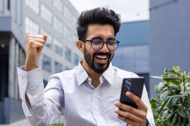 Gözlüklü neşeli erkek, zaferle havayı yumrukluyor. Ofis binasının dışındaki cep telefonundan aldığı haberden memnun..
