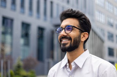 Şehirde güneşli bir günün tadını çıkaran sakallı, gözlüklü ve beyaz gömlekli iyimser bir yetişkin. Sıradan şehir profesyoneli..